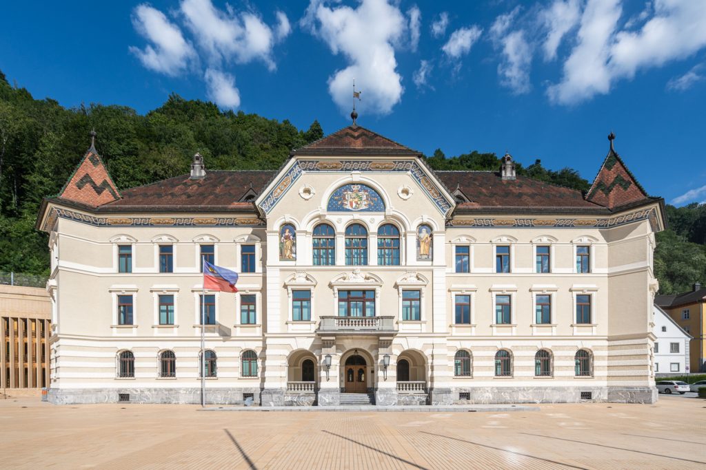 Frontansicht des historischen Regierungsgebäudes in Vaduz mit Flagge und verziertem Giebel.