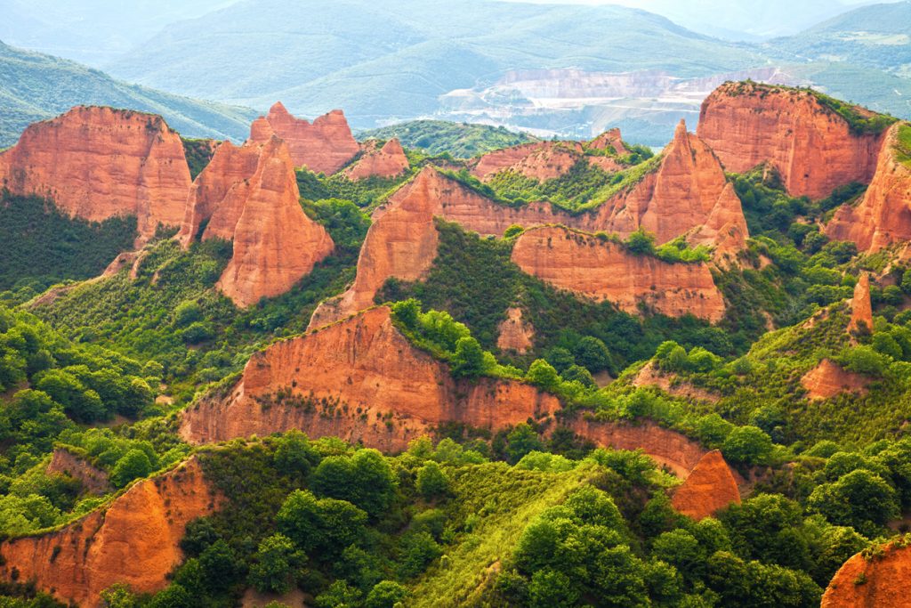Rostrote Hügel und Zacken von Las Médulas heben sich zwischen dichtem Grün und fernen Bergen ab.