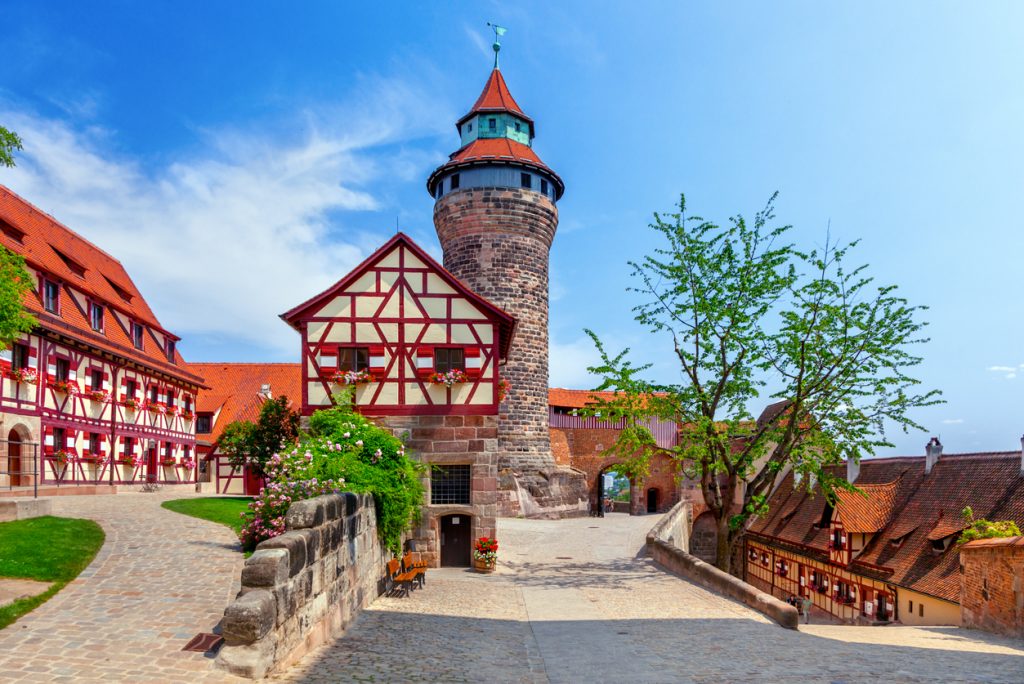 Innenhof der Nürnberger Kaiserburg mit rot-weißem Fachwerkhaus und rundem Sinwellturm unter blauem Himmel.