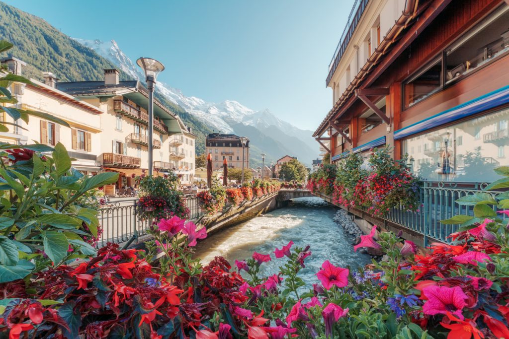 Ufer in Chamonix mit üppigen Blumen, Brücke und Alpenkulisse.