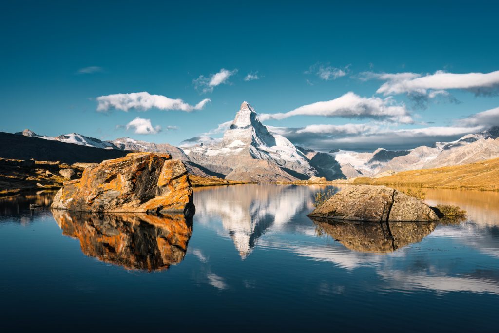Das Matterhorn spiegelt sich im ruhigen Bergsee mit rötlichen Felsblöcken im Vordergrund.