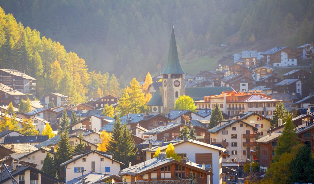 Ortskirche mit Spitzdach zwischen Häusern und gelb leuchtenden Bäumen in Zermatt.