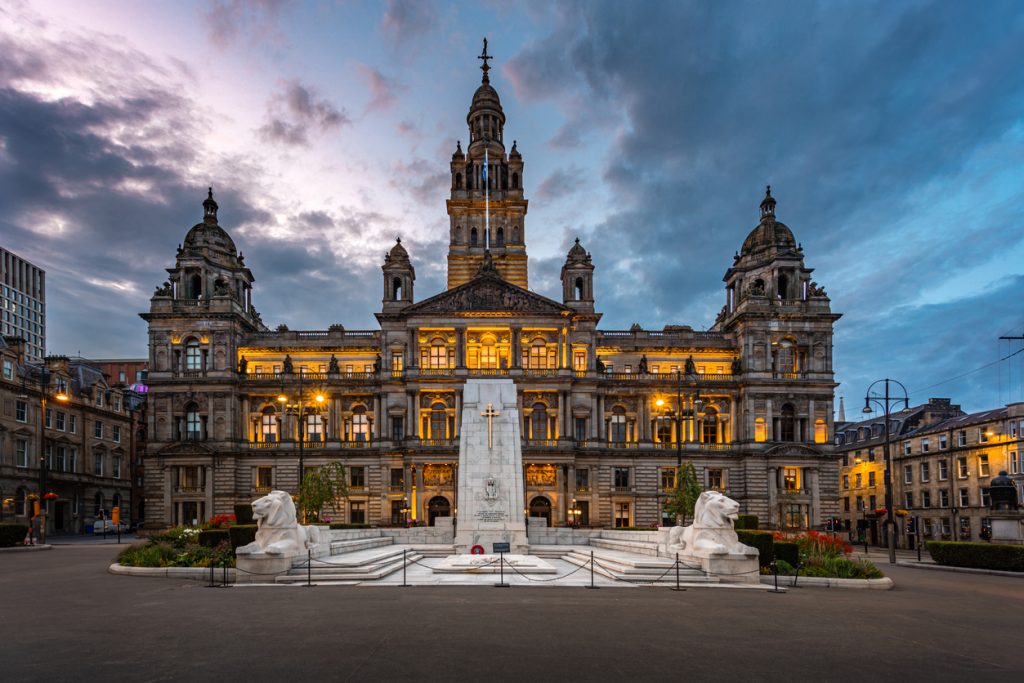Historisches Rathaus mit beleuchteter Fassade und Kriegerdenkmal bei blauem Abendhimmel in Glasgow.