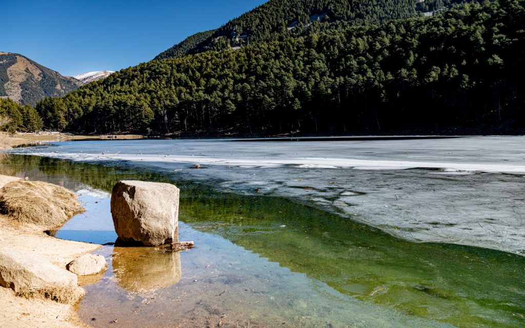 Ufer eines halb zugefrorenen Sees mit klaren Wasserschichten und dunklem Nadelwald.