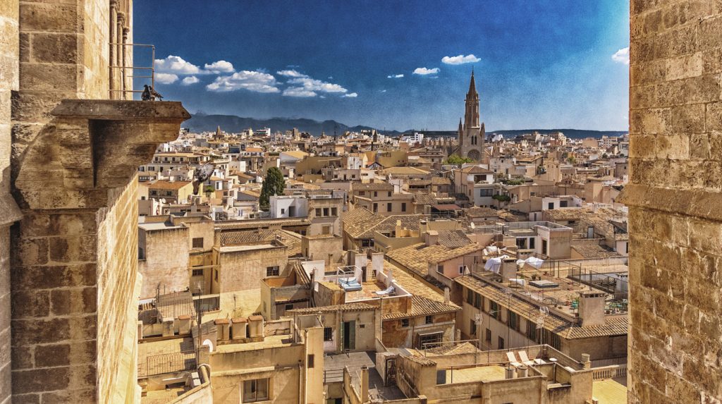 Weitblick über sandfarbene Altstadtdächer mit Kirche und fernen Bergen.
