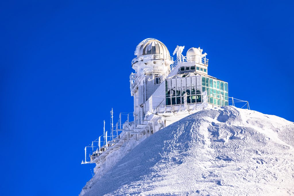 Eisbedecktes Observatorium steht auf einem schneebedeckten Gipfel vor tiefblauem Himmel.