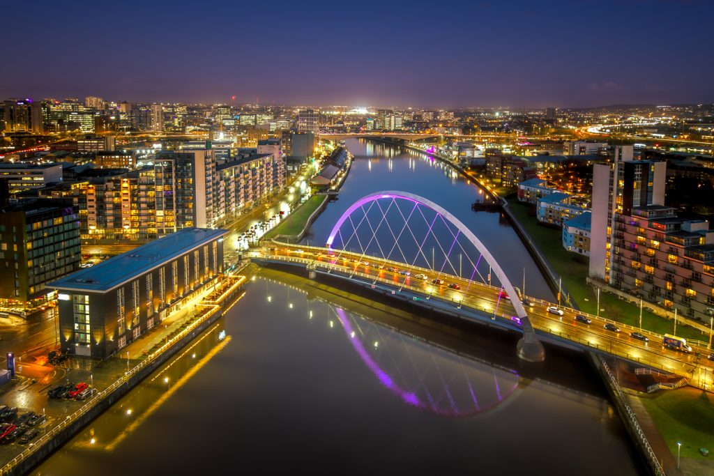 Nachtaufnahme der illuminierten Bogenbrücke und moderner Uferpromenaden in Glasgow.