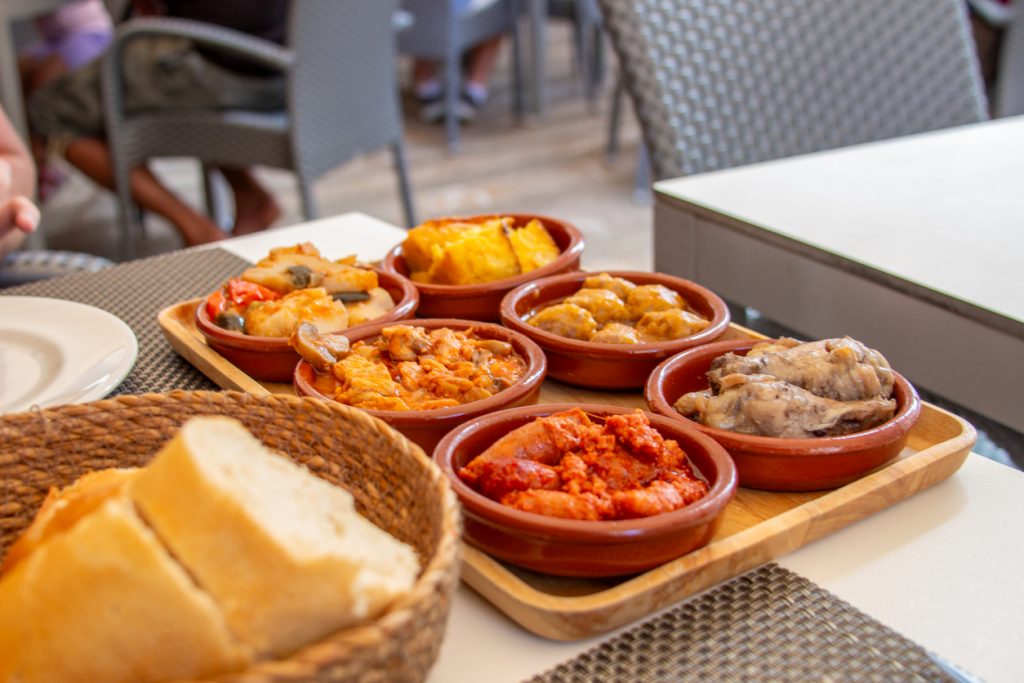Holzbrett mit mehreren kleinen Tontöpfen voller spanischer Tapas neben Brotkorb.
