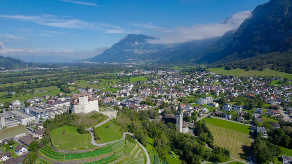 Luftaufnahme von Balzers in Liechtenstein mit Burg Gutenberg, Weinbergen und Bergen im Hintergrund.