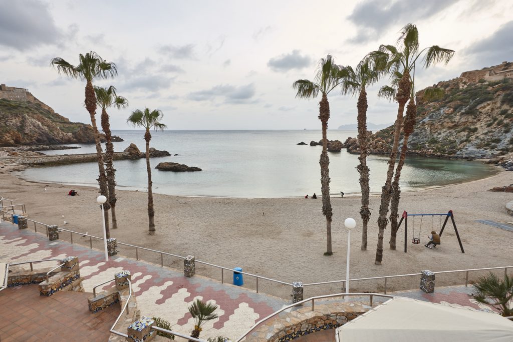 Kleine Sandbucht mit Palmen, ruhigem Wasser, Felsenküste und Promenade.