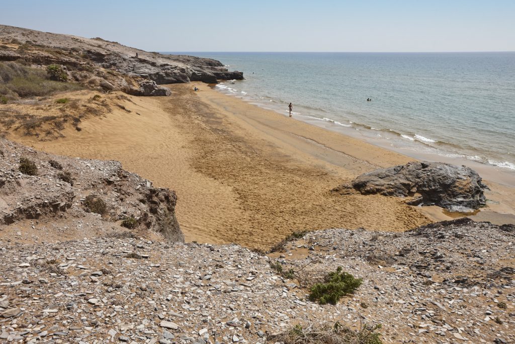 Langer, unverbauter Sandstrand mit Felsköpfen, sanften Wellen und karger Küstenlandschaft