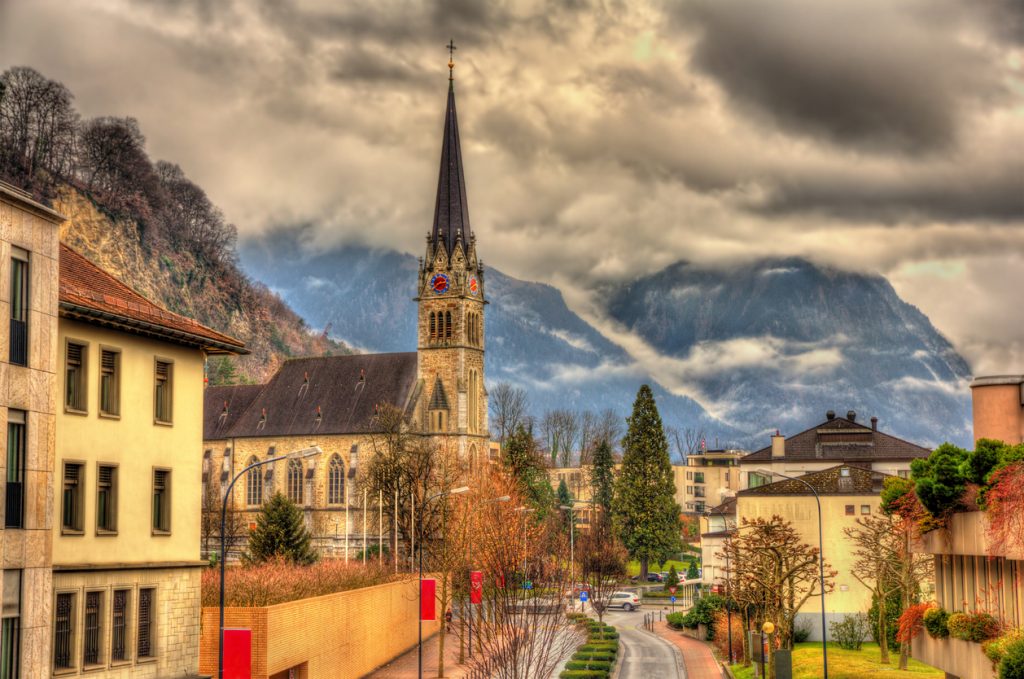 Straßenszene in Vaduz mit der neugotischen St.-Florin-Kathedrale und wolkenverhangenen Bergen.
