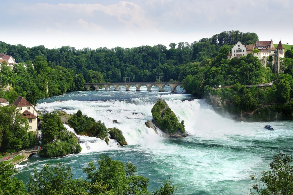 Breiter Wasserfall mit Felsen, Brücke und grünen Ufern am Rhein.
