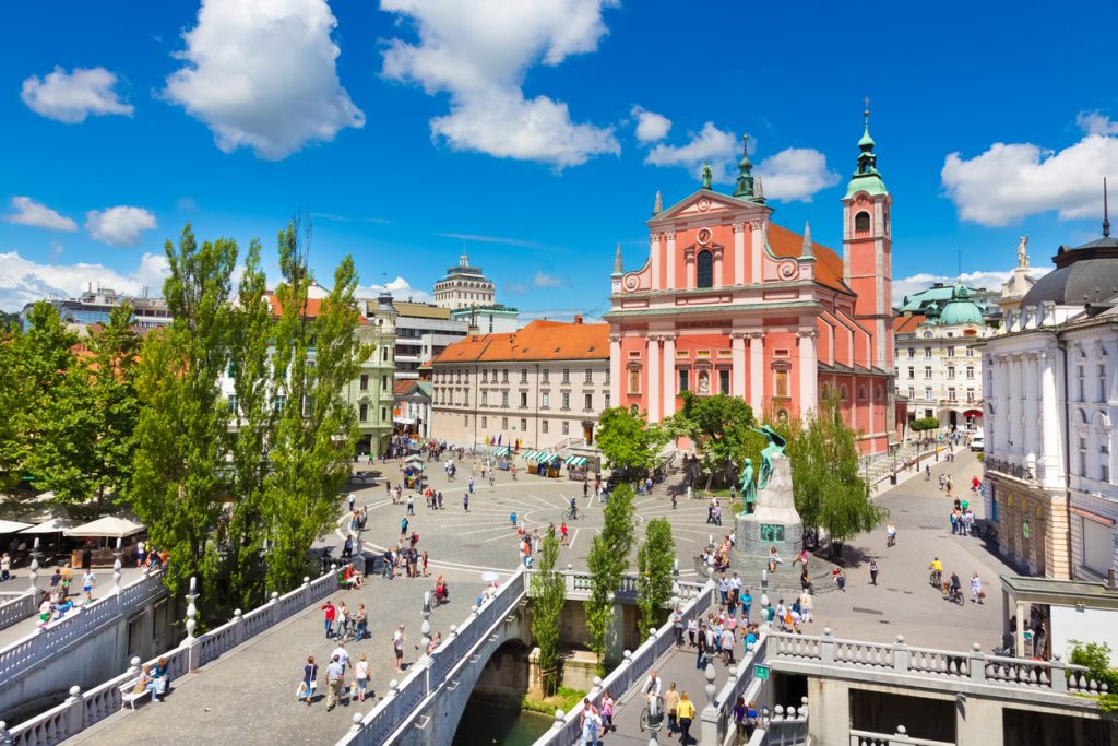 Großer Platz mit rosa Kirche, vielen Menschen und der Dreifachbrücke.