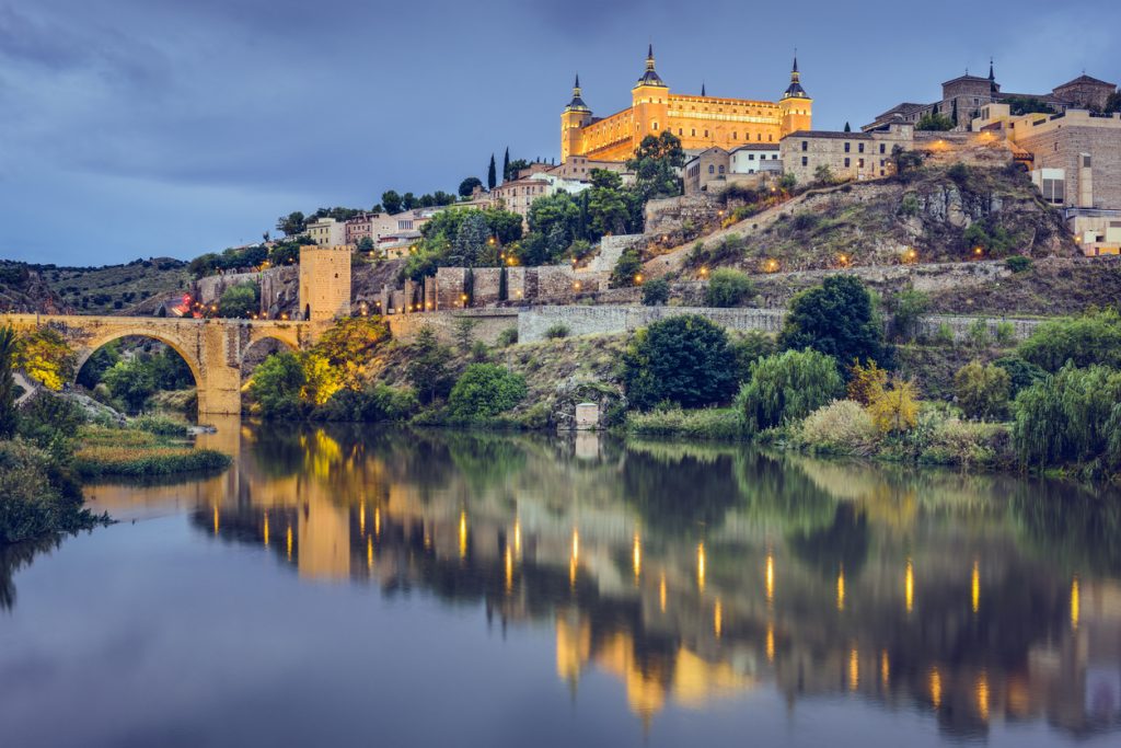 Beleuchteter Alcázar und Stadtmauer spiegeln sich abends im Fluss Tajo.