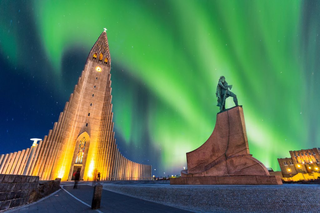 Beleuchtete Hallgrímskirkja in Reykjavík mit Leifur-Eiríksson-Statue vor grünem Polarlicht.