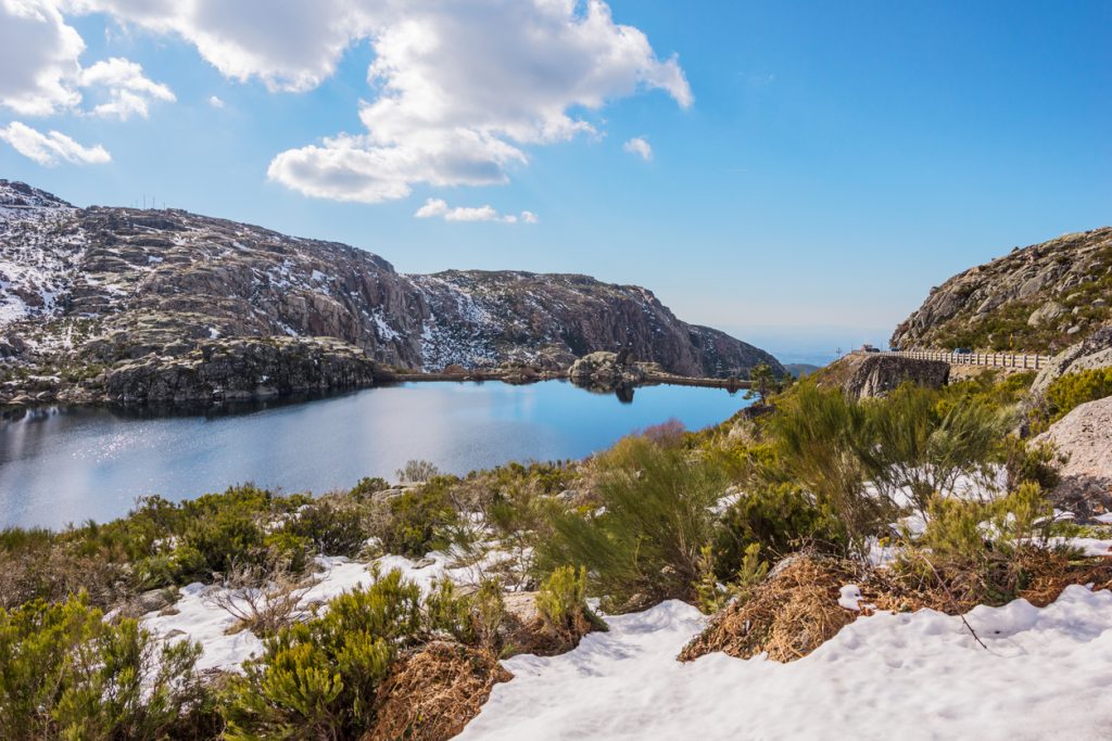 Ein stiller Bergsee liegt zwischen felsigen Hängen mit Schneeresten unter blauem Himmel in Portugal.