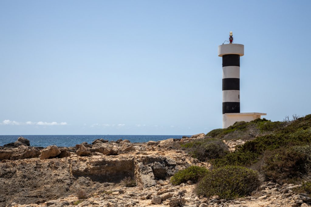 Schwarz-weiß gestreifter Leuchtturm Far de s’Estalella steht auf einer felsigen Küste am Meer.