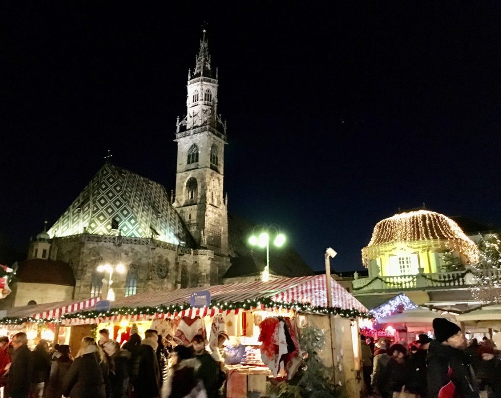 Menschen schlendern abends über einen festlich beleuchteten Weihnachtsmarkt vor dem Dom von Bozen.