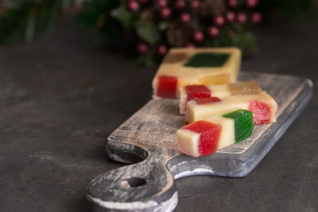 In Streifen geschnittener Turrón mit roten, grünen und gelben Fruchtstückchen liegt auf einem kleinen Holzbrett.