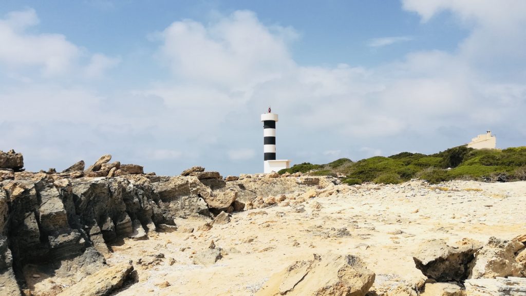 In einer felsigen Küstenlandschaft steht der Far de s’Estalella als kleiner Leuchtturm unter leicht bewölktem Himmel.