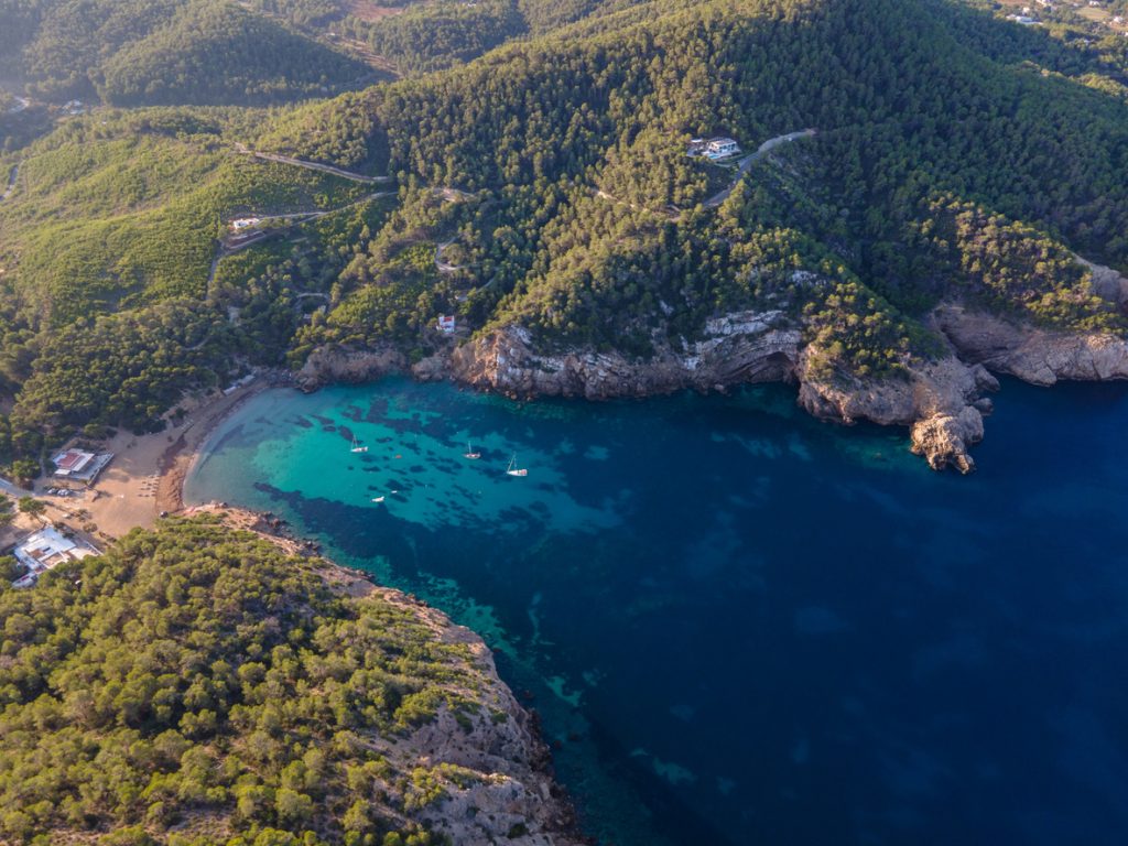 Vogelperspektive auf die Cala Benirrás mit tiefblauer Bucht, grünen Hügeln und kleinen Booten im Wasser.
