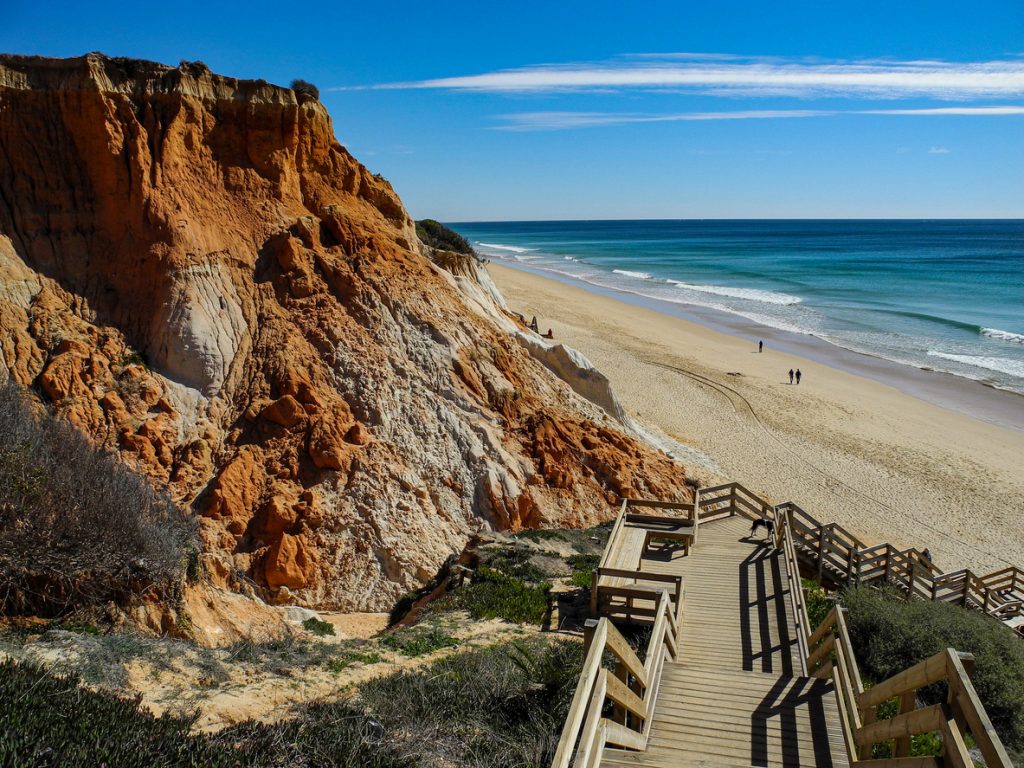 Ein Holzsteg führt an roten Steilklippen hinunter zur Praia da Falésia an der Algarve in Portugal.
