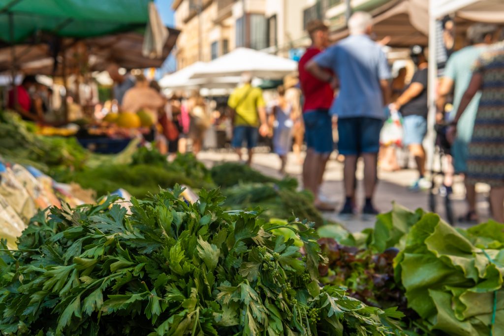 Bündel mit frischen Kräutern und Salaten liegen im Vordergrund, während Marktbesucher und Stände in Santanyí unscharf im Hintergrund zu sehen sind.