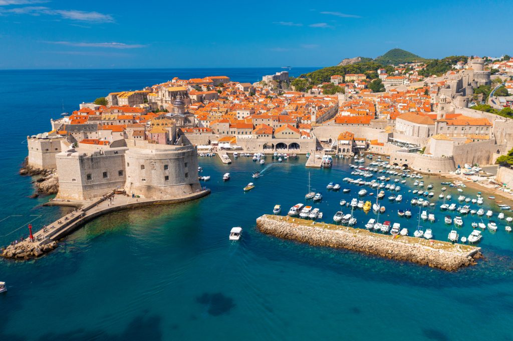 Blick auf Dubrovnik mit Festungsmauern, orangefarbenen Dächern und Booten im türkisblauen Hafen.