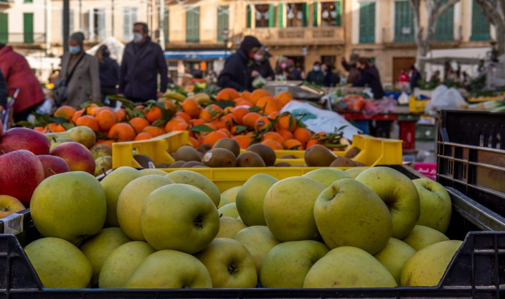 Obstkisten mit grünen Äpfeln und Orangen auf dem Wochenmarkt von Llucmajor, im Hintergrund Menschen mit Einkaufstaschen.
