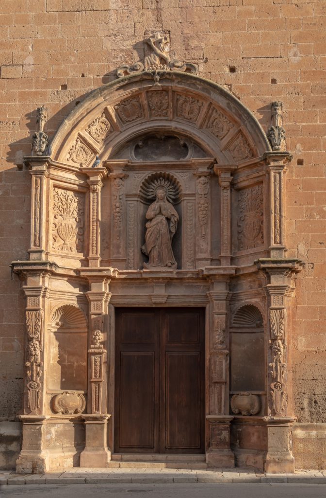 Aufwendig verziertes Sandsteinportal einer Kirche in Llucmajor mit Heiligenstatue über einer dunklen Holztür.