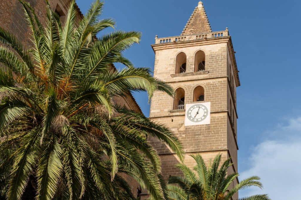 Steinener Kirchturm mit Uhr in Porreres auf Mallorca, davor große Palmen unter blauem Himmel.