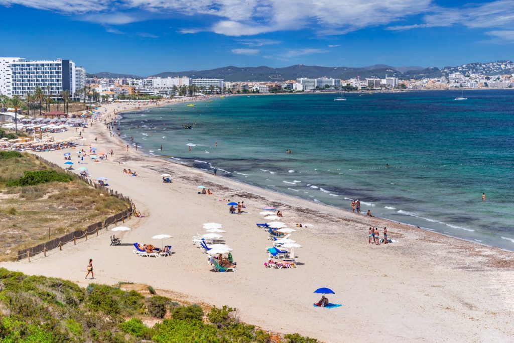 Blick entlang der Playa d’en Bossa mit hellem Sand, Badegästen, Sonnenschirmen und Hotels im Hintergrund.