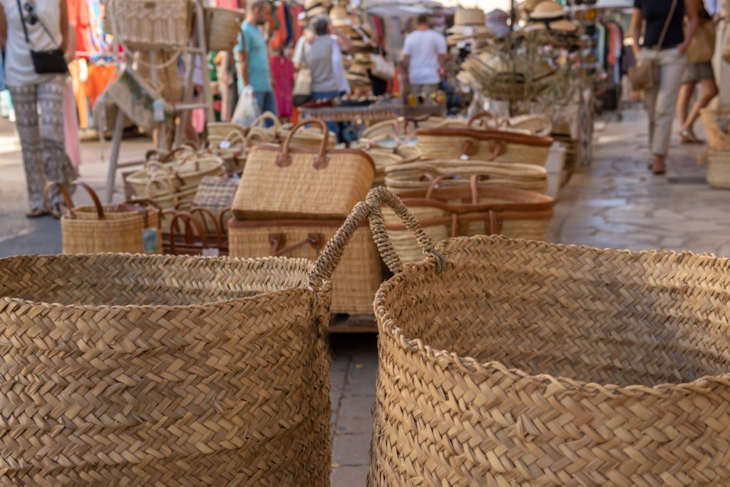 Zwei große geflochtene Körbe im Vordergrund, dahinter zahlreiche Stroh- und Markttaschen sowie schlendernde Besucher auf dem Markt von Santanyí.