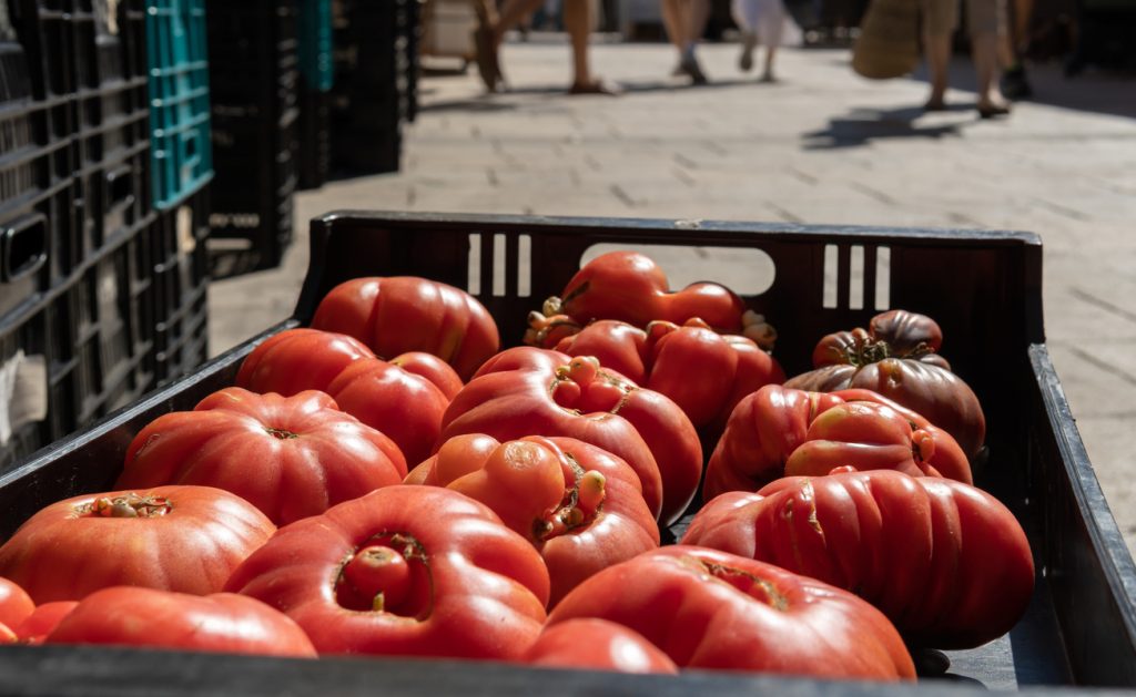 Eine schwarze Kiste voller großer, gerippter Tomaten steht auf dem Boden, im Hintergrund verschwimmen Marktstände und Beine der Besucher in Santanyí.