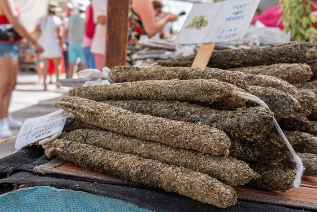 Mehrere lange, mit getrockneten Kräutern ummantelte Würste sind auf einem Marktstand gestapelt, dahinter Menschen und Schilder in Santanyí.