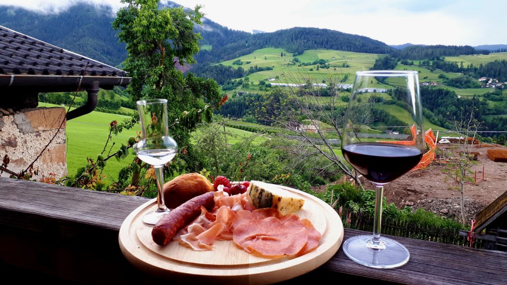 Brett mit Wurst, Schinken, Käse, Brot und zwei Gläsern Schnaps und Rotwein vor Berglandschaft.