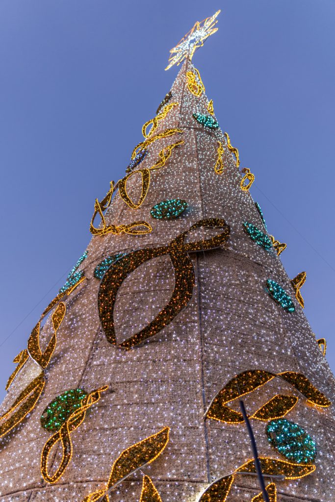 Hoher, kegelförmiger Weihnachtsbaum aus tausenden Lichtern auf einem Weihnachtsmarkt auf Mallorca, von unten gegen den blauen Abendhimmel fotografiert.