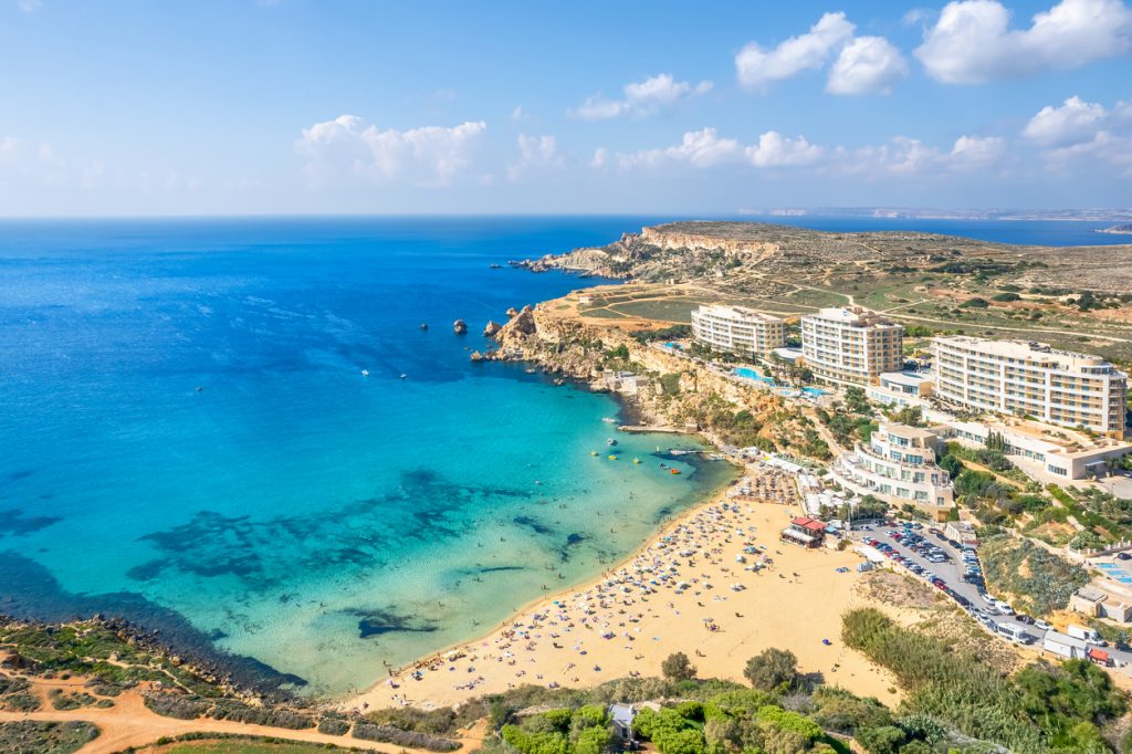 Luftaufnahme der Paradise Bay auf Malta mit goldenem Sandstrand, türkisblauem Meer und Hotels auf den Klippen.