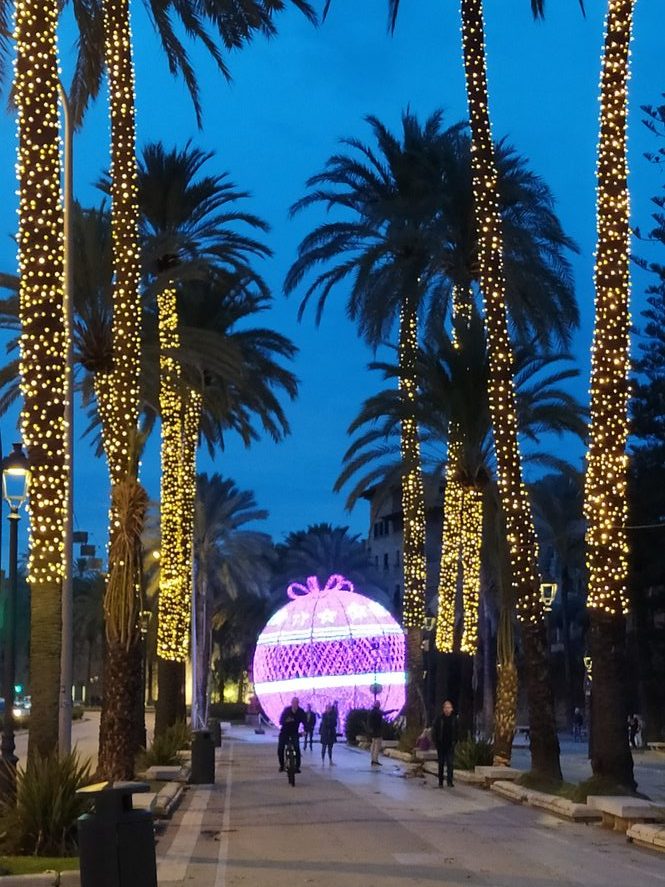 Fußgänger und Radfahrer passieren eine riesige, lila leuchtende Weihnachtskugel am Ende einer mit Lichterketten geschmückten Palmenallee auf Mallorca.