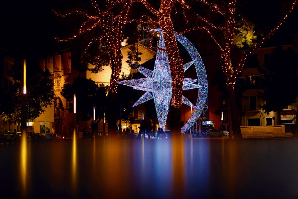 Großes, bunt leuchtendes Sternkunstwerk zwischen Bäumen mit Lichterketten auf einem Weihnachtsmarkt auf Mallorca.