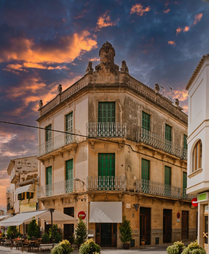 Eckhaus mit grünen Fensterläden und Balkonen in Llucmajor vor einem dramatisch orange-violetten Abendhimmel.