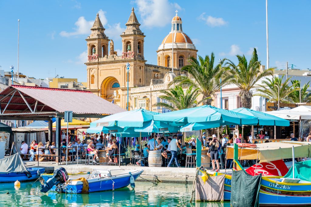 Hafen von Marsaxlokk auf Malta mit bunten Booten, Restaurants und Kirche.