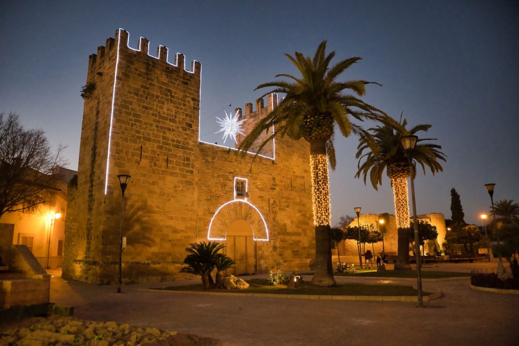 Mittelalterliches Stadttor mit beleuchteten Konturen und umwickelten Palmenstämmen auf einem Weihnachtsmarkt in Alcúdia auf Mallorca.
