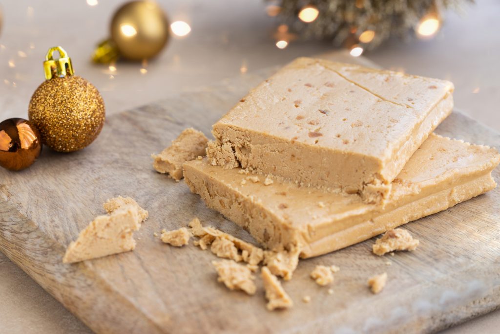 Ein angeschnittener Block Turrón liegt auf einem Holzbrett, darum herum Krümel und goldene Weihnachtskugeln.