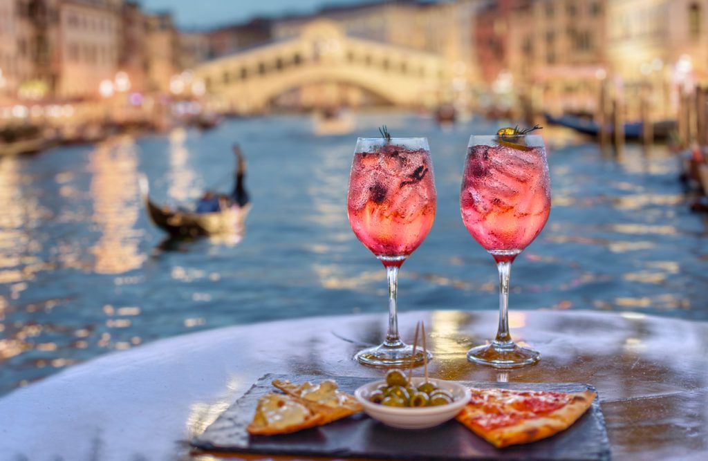 Zwei Gläser Aperitif und Snacks vor dem verschwommenen Hintergrund des Canal Grande und der Rialtobrücke in Venedig.