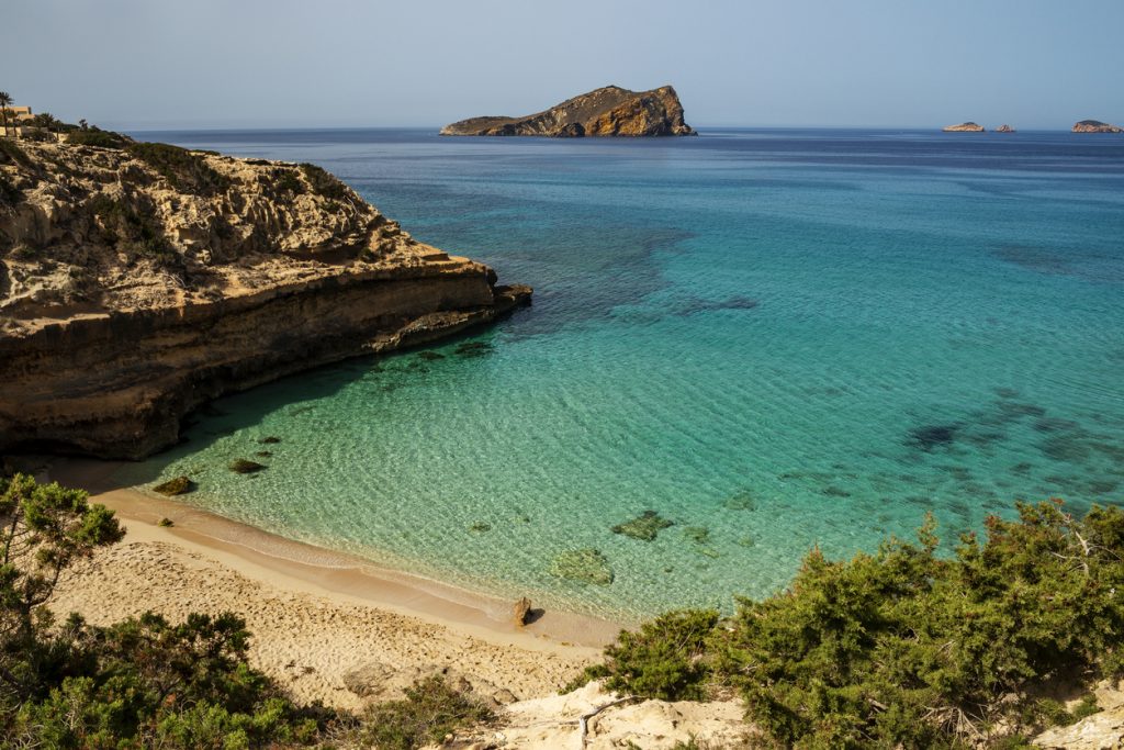 Blick auf die Cala Comte mit heller Sandbucht, Felsenklippen und türkisblauem, klarem Wasser.