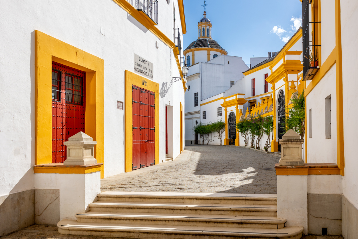 Helle Häuser mit gelben Akzenten und roten Türen bilden eine ruhige Gasse in der Altstadt von Sevilla.