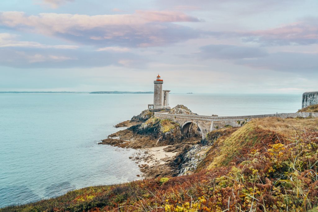 Ein Leuchtturm steht auf einer Felsklippe am Meer in der Bretagne und ist über einen schmalen Damm erreichbar.