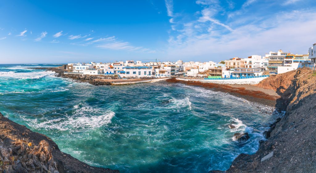 Weißes Dorf an der Felsküste, darunter wirbelt das Meer in einer Bucht auf Fuerteventura.